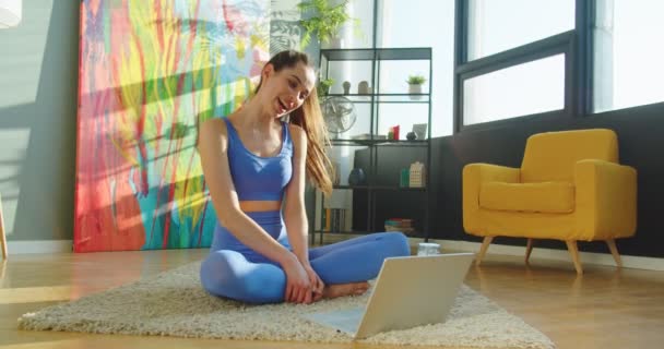 Кавказька щаслива молода спортсменка в спортивній манері сидить на килимовій підлозі розмахуючи рукою і розмовляє по відео на ноутбуку з посмішкою на обличчі. Радісна жінка спілкується на комп "ютері через Інтернет — стокове відео