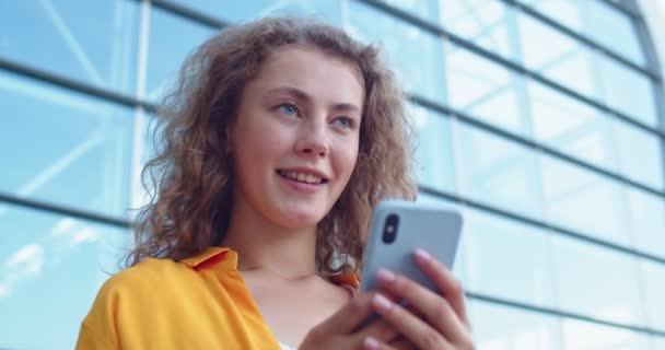 Close up of Caucasian young smiling cheerful woman standing outdoors in city and texting sending message on smartphone with happy face. Beautiful female surfing social network on cellphone online — Stock Video