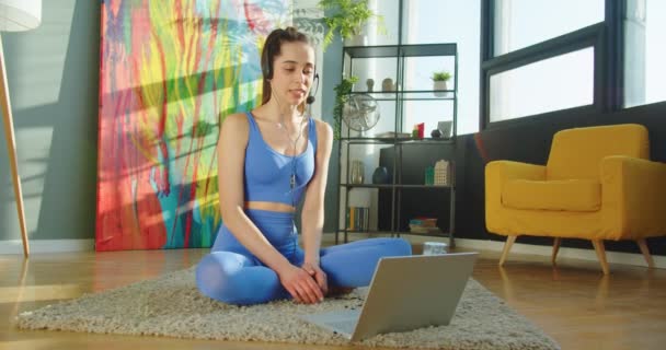 カーペットの床に座って、ラップトップ上のオンラインビデオ通話でヘッドセットで話すスポーツウェアの白人の陽気な若い美しいスポーツウェアの女性。かなり女性のフィットネスインストラクタービデオオートキャストオンコンピュータ — ストック動画