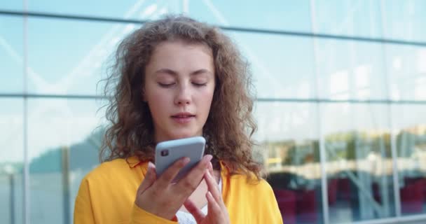 Primer plano retrato de la joven mujer caucásica feliz con el pelo rizado de pie al aire libre y la grabación de mensaje de voz en el teléfono inteligente. Bonita mujer usando un celular en la ciudad en la calle. Concepto de estilo urbano — Vídeos de Stock