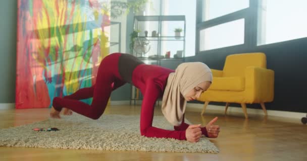 Jovem mulher esportiva muçulmana bonita treinando em casa, de pé na prancha, construindo núcleo forte. Muito focado e motivado feminino fazendo exercício na sala de estar em quarentena, esporte, conceito de treino — Vídeo de Stock