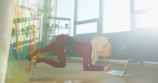 Genç müslüman bir kadın dizüstü bilgisayarda video izleyip evde tahta egzersizi yapıyor. Kilo kaybı için zindelik, kalça kasları, vücut geliştirme, güçlü çekirdek. Çevrimiçi eğitim kavramını kullanarak evden spora geçiş — Stok video