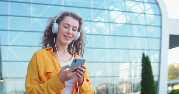 Detailní portrét šťastné mladé krásné bělošky kráčející venku na ulici a poslouchající hudbu ve sluchátkách smskování na smartphonu. Žena se usmívá procházení on-line na mobilu — Stock video