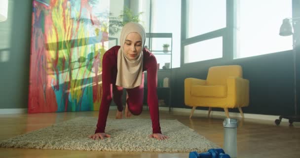 Bastante musulmana feliz hembra haciendo ejercicio de pie en tablón en habitación soleada moderna. Mujer hermosa joven en ropa deportiva que tiene entrenamiento en casa, fitness, estilo de vida deportivo, cuerpo fuerte y núcleo — Vídeos de Stock