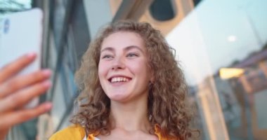 Genç, neşeli, beyaz bir kadının, sokakta dikilip, akıllı telefonuyla görüntülü sohbet ederken cep telefonu iletişim kavramında görüntülü konuşma yapan neşeli bir kadının portresini yakın çekim yapın.