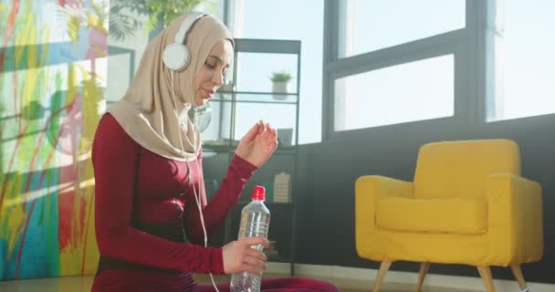 Vacker ung glad muslimsk kvinna lyssnar på musik i hörlurar och dricksvatten efter träning sitter på golvet i moderna rum i huset. Kvinna njuter av musik i soligt hem, fritid koncept — Stockvideo