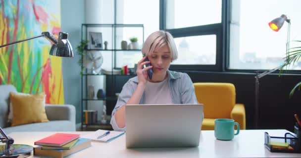 Portret szczęśliwej młodej białej kobiety mówiącej na smartfonie i piszącej na laptopie siedząc przy biurku w gabinecie. Praca na odległość od domu. Piękna uczennica w domu — Wideo stockowe