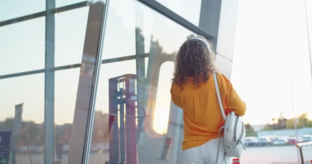 Back view of Caucasian female walking alone outdoors listening favorite music in headphones hears someone calling her turning around and waving hand while smiling in positive mood Walk in city concept — Stock Video