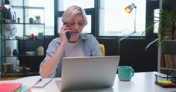 Egy csinos, fiatal, szőke, kaukázusi, örömteli nő portréja, aki mobilon telefonál és laptopon gépel, miközben az asztalnál ül a szekrényszobában. Távolsági munka otthonról. Szabadidős koncepció — Stock videók