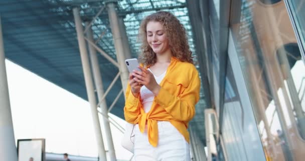 Visão inferior da mulher caucasiana feliz muito encaracolado digitando no celular na rua ao ar livre. Joyful bela jovem internet surf feminino usando aplicativo de rede social na cidade. Conceito de lazer — Vídeo de Stock