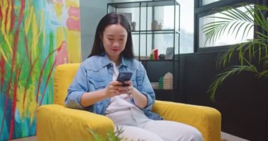 Asyalı mutlu, gülümseyen, sarı koltukta oturan, evinde dinlenen ve sosyal ağ uygulaması kullanarak akıllı telefondan mesaj atan genç bir kadın. Güzel, neşeli bir kadın cep telefonuyla internette geziniyor.
