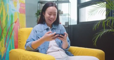 Asyalı neşeli genç kadın evde sarı koltukta oturuyor ve sosyal ağ uygulaması kullanarak akıllı telefondan video izliyor ve gülüyor. Güzel, neşeli kadın cep telefonuyla internette geziniyor.