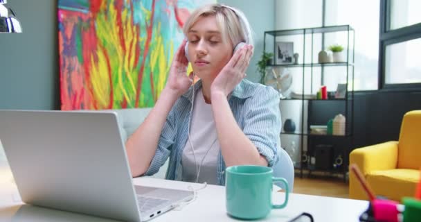 Retrato de mensagens de texto femininas caucasianas jovens navegando on-line no laptop e desfrutando de música em fones de ouvido sentados à mesa no armário do quarto. Aprendizagem estudantil, trabalho à distância, casa, escritório, conceito indoor — Vídeo de Stock