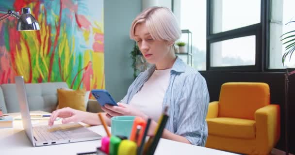 Portrét krásné bělošky, která studuje u stolu na pokoji a píše na notebooku a SMSku na mobilu. Hezké mladé blondýny žena pracující na počítači ve skříni doma — Stock video