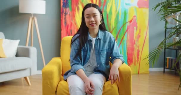 Zamknij portret radosnej młodej Azjatki siedzącej na żółtym krześle w salonie, patrząc w kamerę i uśmiechając się w dobrym nastroju. Piękna szczęśliwa kobieta z uśmiechem na twarzy. Koncepcja czasu wolnego — Wideo stockowe