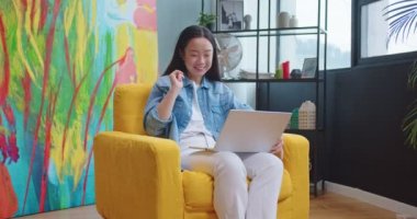 Mutlu Asyalı kadın dizüstü bilgisayarla video sohbeti oturma odasında oturuyor, el sallıyor ve gülümsüyor. Genç, neşeli, güzel bir bayan dairede bilgisayardan video görüşmesi yapıyor.