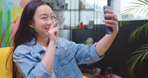 Close up of happy Asian woman videochatting on smartphone sitting in living room in chair and waving hand and smiling Young joyful pretty female in apartment speaking on online video call on mobile phone — Stock video