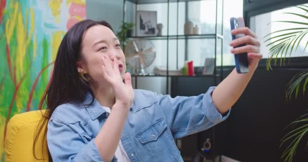 Κοντινό πλάνο της Ασιάτισσας χαρούμενης νεαρής γυναίκας που έχει τηλεδιάσκεψη στο smartphone κάθεται στο σαλόνι και κουνώντας το χέρι με χαμόγελο στο πρόσωπο. Χαρούμενη γυναίκα που μιλάει σε απευθείας σύνδεση βιντεοκλήση στο κινητό στο σπίτι — Αρχείο Βίντεο