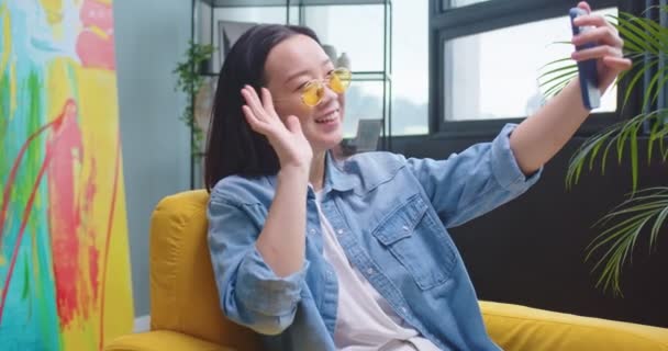 Asyalı, oldukça neşeli, mutlu, sarı gözlüklü bir kadının portresi modern bir odada oturmuş akıllı telefondan sohbet ediyor. Günlük için kadın kayıt videosu, boş zaman konsepti — Stok video