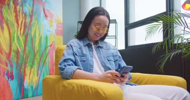 Belle asiatique jeune femme heureuse gaie dans des lunettes jaunes assis dans une chaise dans une chambre moderne à la maison et tapant navigation en ligne sur smartphone. Message texte féminin sur téléphone portable, concept de loisirs — Video