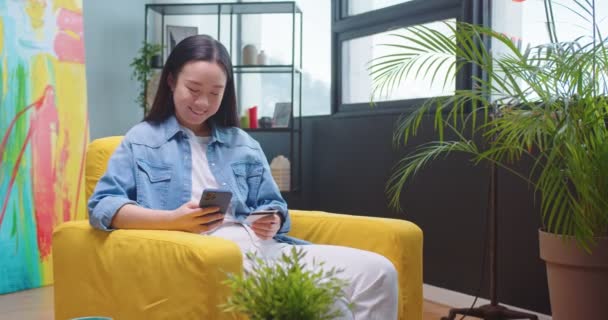Asijské šťastný mladá krásná žena sedí v obývacím pokoji ve žlutém křesle a psaní na smartphone nákup on-line na internetu pomocí kreditní karty platit za nákup. Elektronický obchod, nákupní koncept — Stock video