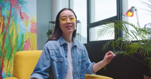 Portrait d'une jeune femme asiatique heureuse avec le sourire sur le visage assis dans une chaise jaune dans une pièce moderne, regardant la caméra et enlevant des lunettes élégantes jaunes. Femme joyeuse posant à la caméra à la maison — Video