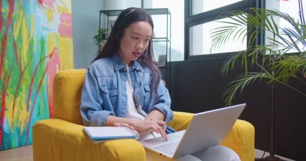 Retrato de una joven asiática estudiante bastante femenina que estudia teniendo una lección en línea en un portátil hablando con auriculares y escribiendo en un cuaderno. Mujer trabajando desde casa teniendo videoconferencia en computadora — Vídeo de stock