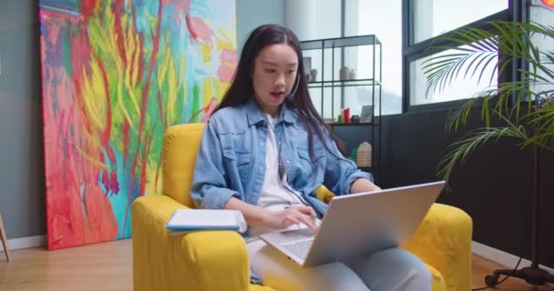 Asiática bela jovem sentada na cadeira amarela no aconchegante armário do quarto e falando sobre chamada de vídeo on-line no laptop falando no fone de ouvido e escrevendo no planejador Estudando on-line, e-learning, conceito de trabalho — Vídeo de Stock