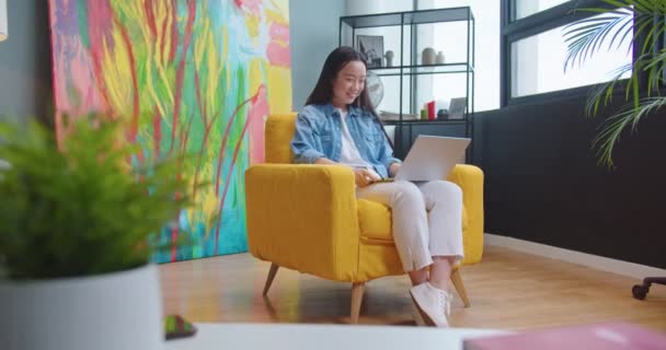 Joyeux asiatique jeune femme vidéochat sur ordinateur portable reposant dans le salon dans une chaise jaune et souriant. Joyeux beau femelle parlant sur appel vidéo en ligne sur ordinateur à la maison intérieur, concept de loisirs — Video