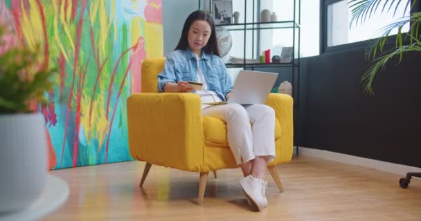 Asiatique heureux jeune belle femme assise dans le salon dans une chaise jaune et tapant sur ordinateur portable achat en ligne sur Internet en utilisant la carte de crédit payer pour l'achat. E-commerce, concept de shopping — Video