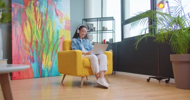 Joyeux positif asiatique jeune femme écouter de la musique dans les écouteurs et dactylographier sur Internet de recherche d'ordinateur portable tout en étant assis sur une chaise dans une chambre confortable moderne. loisirs, travail à distance, étude en ligne — Video
