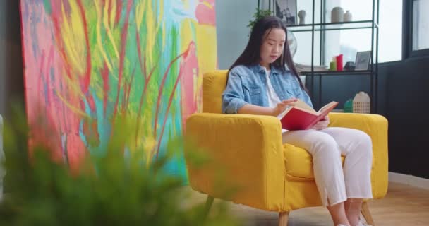 Asyalı mutlu genç kadın modern oturma odasındaki sarı koltukta oturuyor ve romantizm kitabı okuyor. Güzel kız öğrenci çalışıyor, dinleniyor, hobi, boş zaman, okuyucu konsepti — Stok video