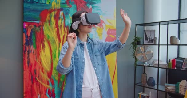 Retrato da jovem asiática feliz em óculos VR usando tecnologia futurista moderna e mãos em movimento no ar em pé no quarto moderno no apartamento. Feminino trabalhando em fone de ouvido de realidade virtual — Vídeo de Stock
