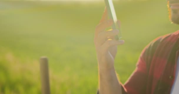 Gehakt beeld van de optimistische blanke boer wetenschapper in geruit shirt chatten via smartphone mobiel tijdens het werken op het veld en glimlachen. Landbouwconcept — Stockvideo