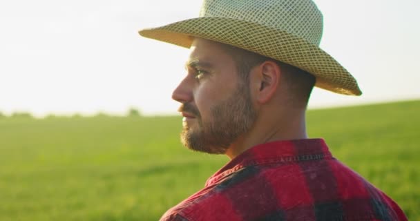 Portrét atraktivního mladého bělocha v klobouku, jak stojí na zelené louce, vesele se usmívá a raduje se, když se dívá na pole. Muž farmář s úsměvem venku v létě — Stock video