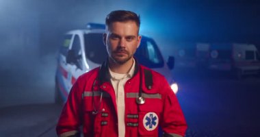 Kameraya bakıp dışarıda dikilen, kırmızı üniformalı beyaz, yakışıklı bir sağlık görevlisi portresi. Arka planda ambulans arabası var. Gece vardiyasında çekici, ciddi bir erkek doktor..