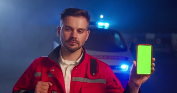 Porträt eines hübschen jungen kaukasischen Sanitäters in roter Uniform, der nachts draußen auf der Straße steht und sein Smartphone mit grünem Bildschirm in der Hand hält. Gut aussehender Mann Arzt zeigt Telefon. Chroma-Schlüssel. — Stockvideo