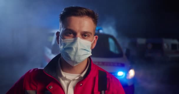 Retrato branco bonito jovem paramédico em máscara médica. Médico masculino atraente no respirador olhando para a câmera. Médico cansado à noite durante a pandemia do covid-19. Carro de ambulância no fundo. — Vídeo de Stock