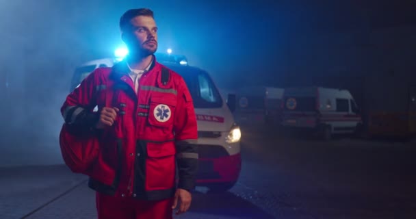 Un bel giovane paramedico caucasico in uniforme rossa che si prepara a lavorare di notte. Auto di ambulanza sullo sfondo. Bell'uomo medico all'aperto con lo zaino. Ritratto. — Video Stock