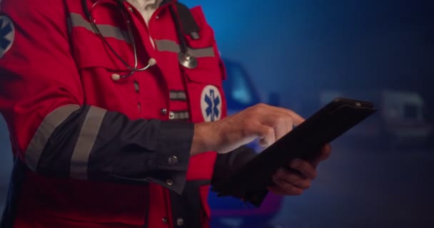 Gros plan de l'ambulancier masculin caucasien en uniforme rouge tapant et tapant sur l'écran de la tablette. Homme remplissant le formulaire médical sur l'appareil la nuit en plein air. Urgence. — Video