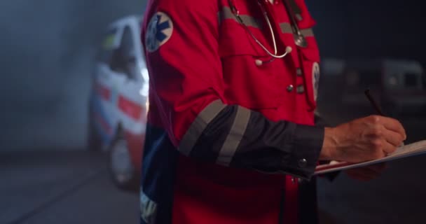 Närbild av kaukasiska manliga paramedicin i röd uniform skrift med penna i hand i mapp med medicinska kort och dokument. Människan medic fylla i medicinsk form på natten utomhus. Nödsituation. — Stockvideo