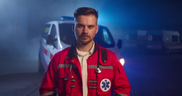 Retrato de caucasiano bonito jovem paramédico em uniforme vermelho olhando para a câmera e de pé ao ar livre. Carro de ambulância no fundo. Médico masculino grave e atraente no turno da noite. — Vídeo de Stock