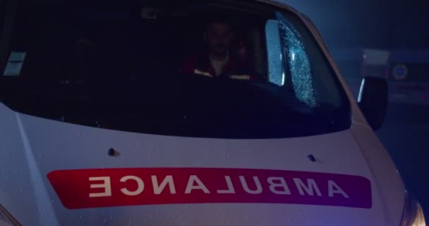 Giovane, affascinante paramedico caucasico in uniforme rossa che guida un'ambulanza, si ferma e esce. Medico maschile stanco che cammina in strada di notte. Chiamata d'emergenza. Lavoro a turni di notte del medico. — Video Stock