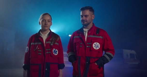 Ritratto di giovane uomo e donna caucasica sorridente paramedici all'ambulanza. Medici maschi e femmine in uniforme rossa che si sorridono a vicenda e alla telecamera. Medici e colleghi di notte. — Video Stock