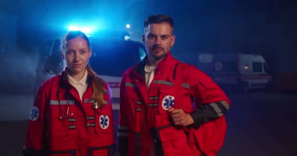 Ritratto di un serio giovane caucasico e paramedico donna all'ambulanza. Medici uomini e donne in uniforme rossa che guardano la macchina fotografica. Medici e colleghi di turno di notte. — Video Stock