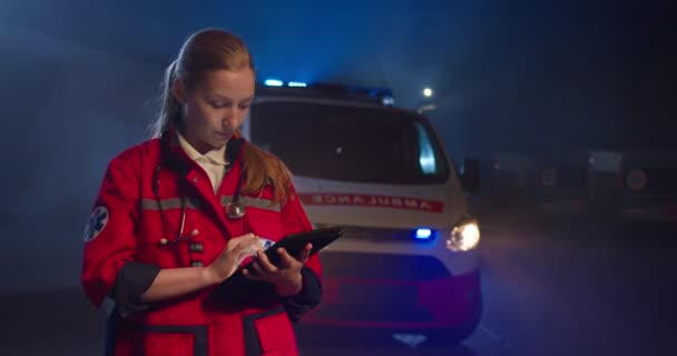 Giovane paramedica caucasica in uniforme rossa che tocca e digita sullo schermo del tablet. Donna medico compilando modulo medico sul dispositivo durante la notte all'aperto. Chiamata ambulanza di emergenza. — Video Stock