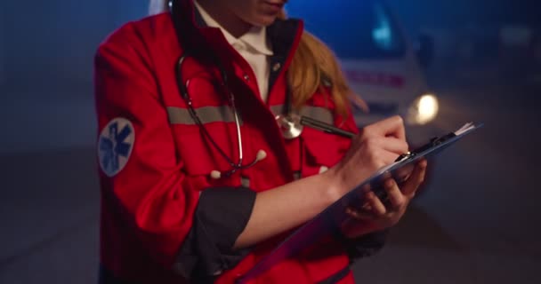 Primer plano de paramédico femenino caucásico en uniforme rojo escrito con pluma en la mano en carpeta con tarjetas médicas y documentos. Mujer médico relleno en forma médica en la noche al aire libre. Emergencia. — Vídeo de stock
