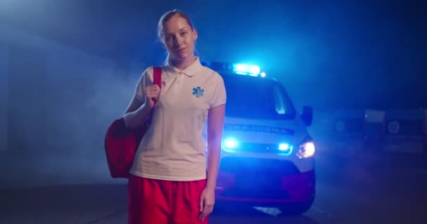 Bionda caucasica giovane paramedica in uniforme che si prepara a lavorare di notte. Auto di ambulanza sullo sfondo. Bella donna medico all'aperto con zaino. Ritratto. — Video Stock