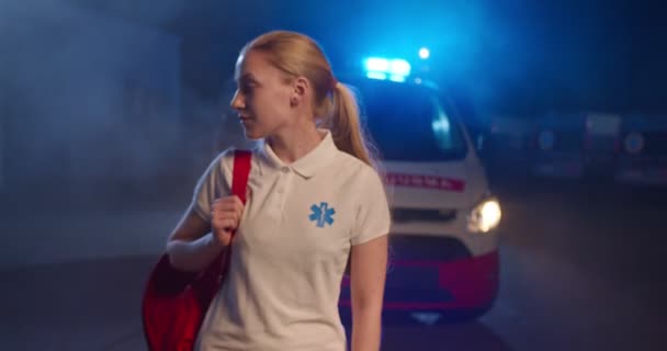 Porträtt av vit blond ung kvinnlig sjukvårdare i uniform redo att arbeta på nattskiftet. Ambulans bil på bakgrunden. Snygg kvinna läkare eller sjuksköterska utomhus med ryggsäck. — Stockvideo