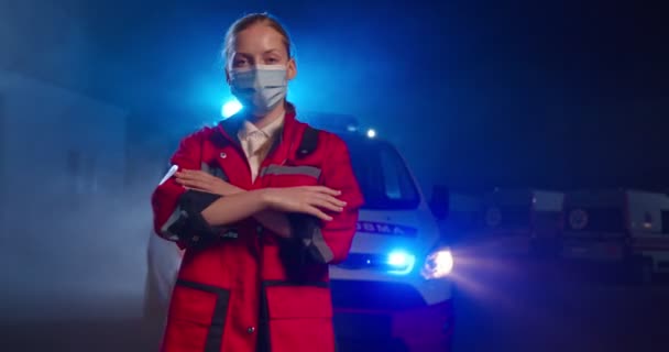 Retrato de paramédico feminino caucasiano em uniforme vermelho e máscara médica olhando para a câmera, cruzando as mãos ao ar livre. Ambulância no fundo. Médica no turno da noite. pandemia de coronavírus. — Vídeo de Stock